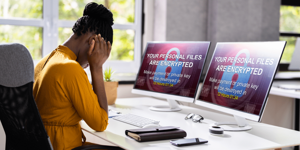 Lady sitting at a desk with two screens holding her head. Ransomware message on both computer screens.